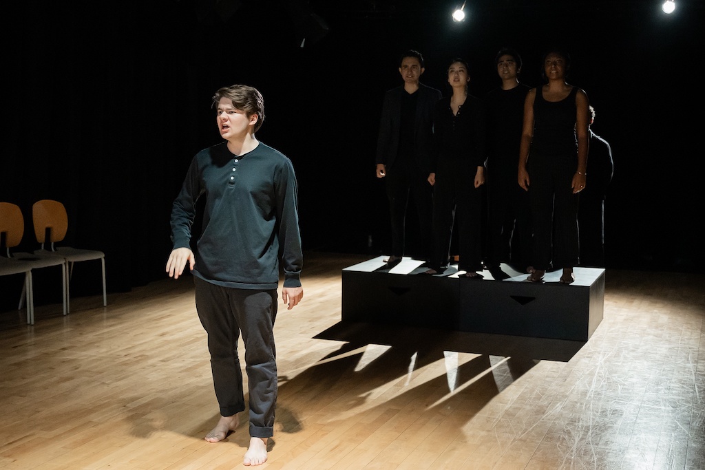 An actor stands on stage in mid-speech while a group of actors stand on a black riser behind him.